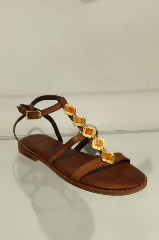 Sandale plate en cuir chocolat ornée de cabochons dorés sur le cou-de-pied. Entièrement en cuir véritable avec première rembourrée pour un confort optimal. Maintien assuré par une sangle tour de cheville ajustable. Petit talon discret pour une allure élégante et intemporelle.