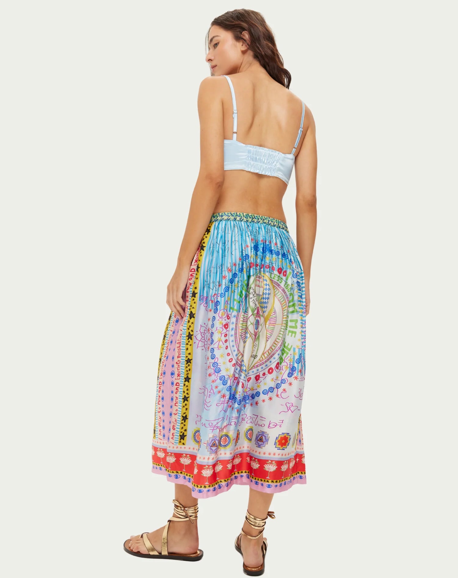 Jupe midi Vanessa aux sept symboles des chakras alignés verticalement dans des couleurs arc-en-ciel éclatantes. Des rayures turquoise en haut se fondent progressivement dans des teintes rose doux et lilas, ornées de mystérieux scripts runiques et de géométrie sacrée. La taille élastique froncée crée du volume et du mouvement fluide, tandis que des bordures mandala colorées en corail, orange, jaune et violet encadrent l'ourlet avec des détails pointillés décoratifs. Les motifs spiralés et symboles spirituels